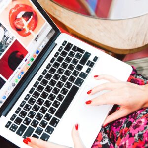 A woman with red nails works on a laptop browsing fashion websites, showcasing online trends.